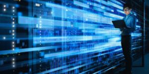 Shot of Asian IT Specialist Using Laptop in Data Center Full of Rack Servers. Concept of High Speed Internet Connections with Blue Visualization Projection of Binary Data Transfer